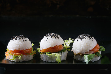Mini rice sushi burgers with smoked salmon, green salad and sauces, black sesame served on black square plate with textile napkin over black background. Modern healthy food