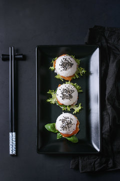 Mini rice sushi burgers with smoked salmon, green salad and sauces, black sesame served on black square plate with chopsticks and textile napkin over black background. Modern healthy food. Top view