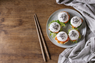 Mini rice sushi burgers with smoked salmon, green salad and sauces, black sesame served on gray plate with wooden chopsticks and textile over wood background. Modern healthy food. Top view with space