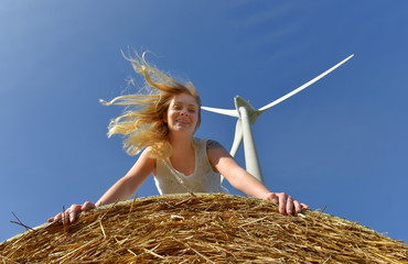 A young blonde girl dressed in a white dress stands on a hay bale with a hugh wind turbine above her.