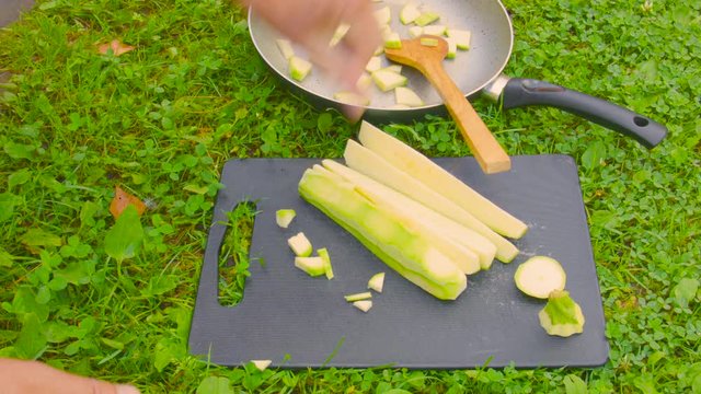 Cook hand chopping zucchini and dropping it on the frying pan laid on the green lawn. Time Lapse Video
