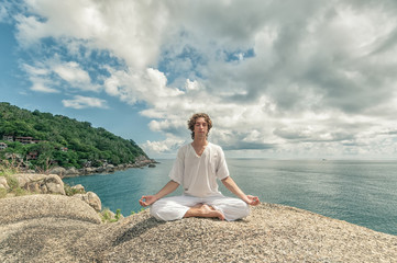 Young man meditating on a high cliff above the sea in the Lotus position - obrazy, fototapety, plakaty
