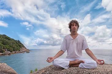 Young man meditating on a high cliff above the sea in the Lotus position - obrazy, fototapety, plakaty