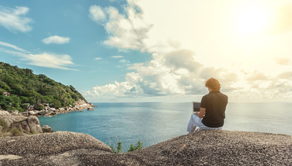 Young man, the freelancer sits on a high cliff over the sea and works remotely - obrazy, fototapety, plakaty