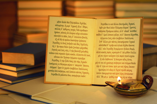 Open Book Illuminated By An Oil Lamp. The Opening Lines Of The Iliad Written In The Ancient Greek Language. A Stack Of Books. On The Lamp, Visible Inscription Aphrodite In The Greek Alphabet.