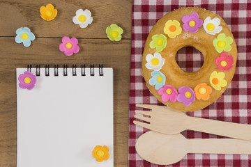 Top view of a fancy donut on wooden table with a spoon, fork and opened notepad