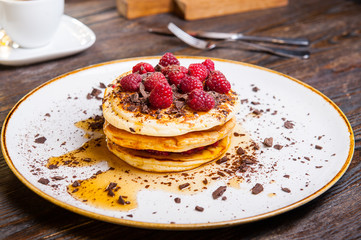 Delicious pancakes with fresh raspberry, chocolate crumbs and maple syrup on the white round plate on the served for breakfast dark wooden table. close-up. selective focus.