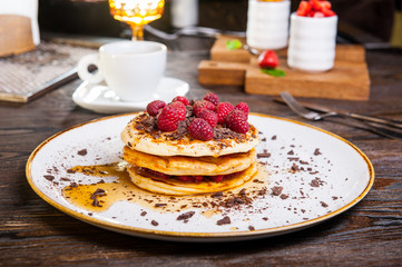 Delicious pancakes with fresh raspberry, chocolate crumbs and maple syrup on the white round plate on the served for breakfast dark wooden table. close-up. selective focus.