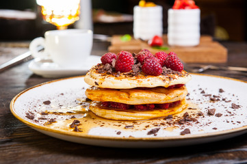 Delicious pancakes with fresh raspberry, chocolate crumbs and maple syrup on the white round plate on the served for breakfast dark wooden table. close-up. selective focus.