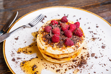 Delicious pancakes with fresh raspberry, chocolate crumbs and maple syrup on the white round plate on the served for breakfast dark wooden table. close-up. selective focus.
