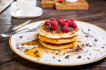 Delicious pancakes with fresh raspberry, chocolate crumbs and maple syrup on the white round plate on the served for breakfast dark wooden table. close-up. selective focus.