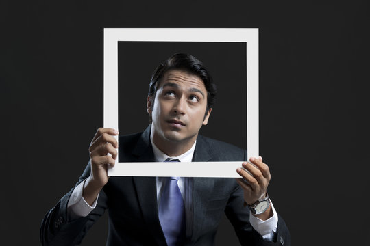 Young businessman looking up while holding picture frame over black background