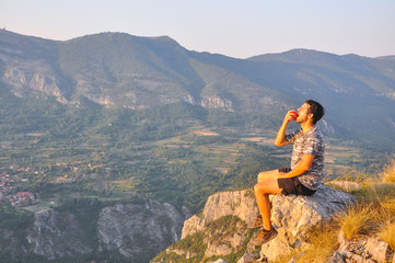 Naklejka premium Young man sits on a cliff edge on the top of mountain with gorgeous view and eats. Dangerous Place. Adrenaline. Hiker on the top