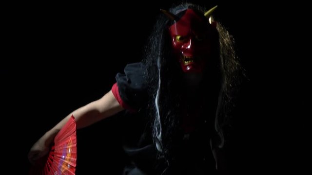 A freak in a mask of a Japanese demon with long hair come ous from darkness with a red fan and hides in the dark