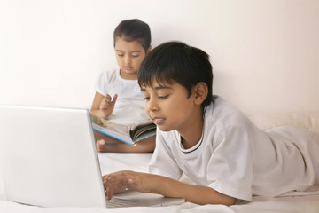 Boy using laptop while girl reading book in bed