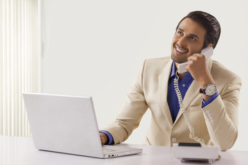 Young businessman talking on telephone while using laptop at office desk