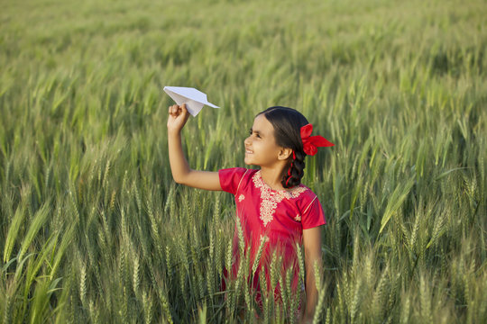 Little Indian girl holding a paper aero plane standing in wheat field 