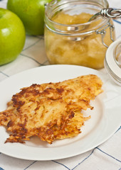 Organic potato fryed, pancake on a round, white plate with a clear, glass jar of homeande applesauce, two ripe, raw, local green apples, white table cloth, blue stripes