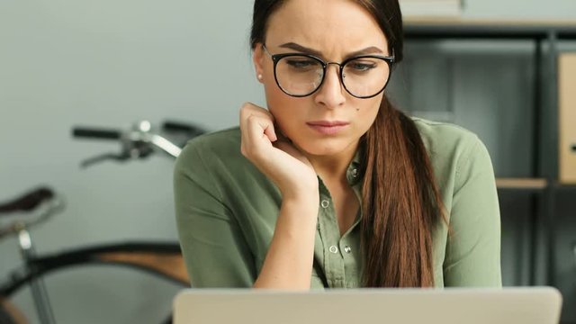 Close up of young atractive woman wearing glasses and working with laptop computer in modern urban office. Woman thinking while looking at the window.