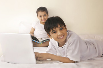 Portrait of boy using laptop with girl reading book in bed