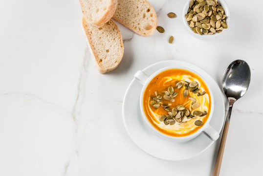 Traditional fall and winter dishes, hot and spicy pumpkin  soup with pumpkin seeds, cream and freshly baked baguette, on white marble table, copy space top view
