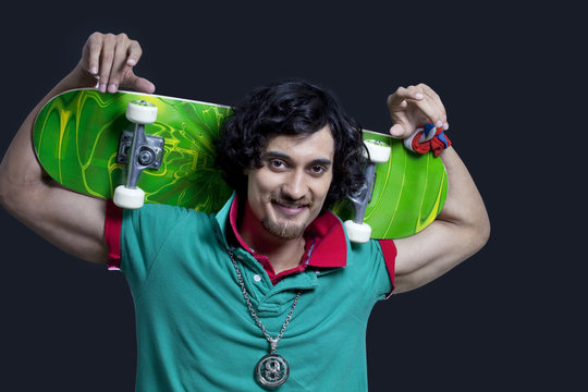 Portrait of young smiling man holding skateboard behind head against black background 