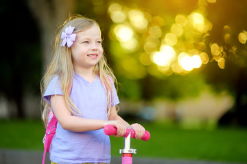 Little child learning to ride a scooter in a city park on sunny summer day. Cute preschooler girl...