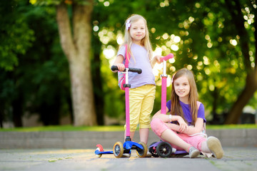 Small children learning to ride scooters in a city park on sunny summer evening. Cute little girls riding rollers. © MNStudio