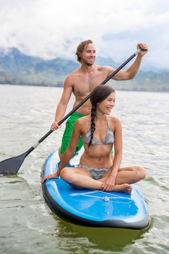 Paddleboard couple having fun paddleboarding together on Hawaii beach vacation summer holidays. Woman sitting on board while man paddles.