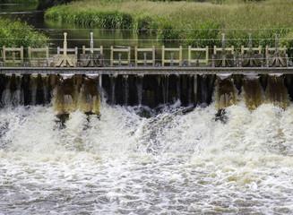 Brown and foamy water flowing through sluice underneath a narrow wooden bridge.