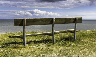 Old empty wooden bench on the beach with a view over the sea. Cloudy sky background.