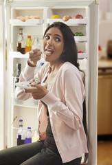 Young woman eating cake in front of open refrigerator 