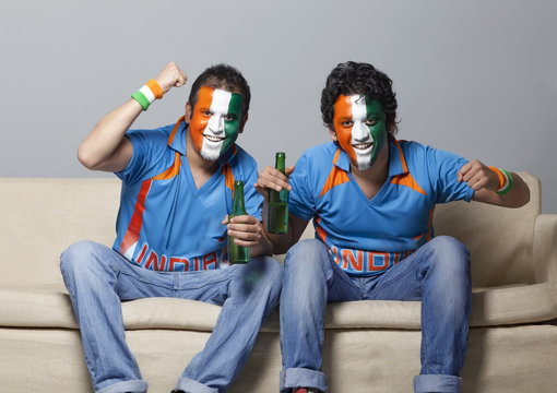 Portrait of male cricket fans with face painted in Indian tricolor cheering while having drink at home 