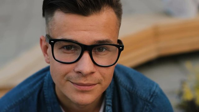 Portrait of young handsome man in the glasses looking to the camera and smiling on the city background. Close up shot.