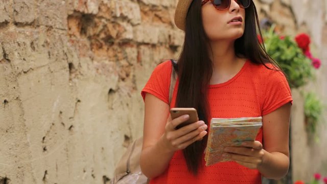 Young attractive woman walking in the old city street. Female traveler using paper map and online maps on smartphone to find interesting places. Close up