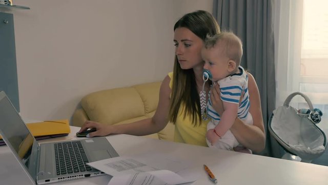 Young mother works on the laptop with baby in arms