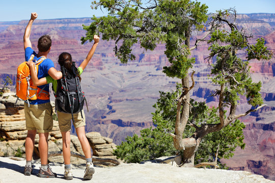 Grand Canyon hikers tourists couple cheering success with arms up happy for hike achievement. Backpackers hiking with backpacks standing at view of famous american landmark, USA trip travel.