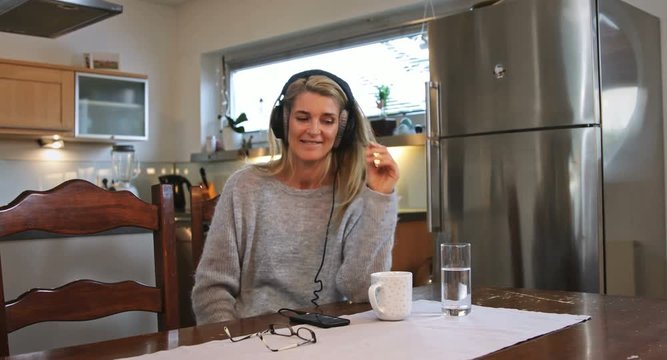 A woman listens to music through headphones. Two cups, a mobile phone, and glasses lay around on the table.