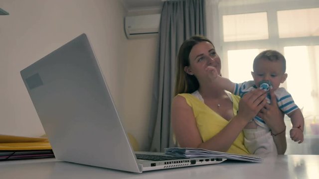 Young mother holds a little son on arms and tries to work at the laptop