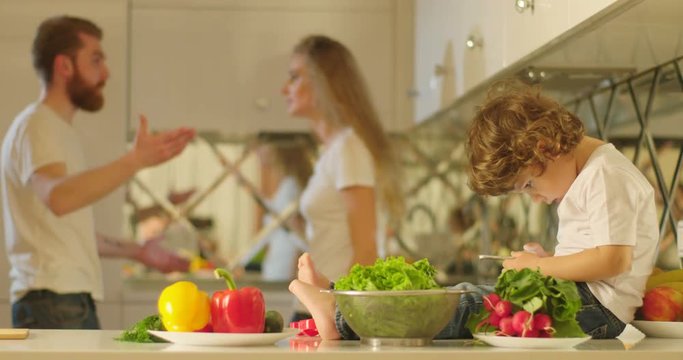 The little boy with curly hair is using the mobile phone while sitting on the kitchen furniture full of different vegetables at the background of the mother is slapping the father.