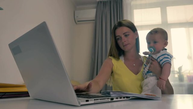 Young mother is working at home with laptop at the table and hold a baby in arms