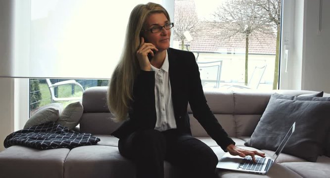 A business woman working from home talks on the phone and looks at her laptop. One window blind is raised and is letting in sunlight into the room.