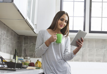 Young woman holding cup of tea and using digital tablet in kitchen