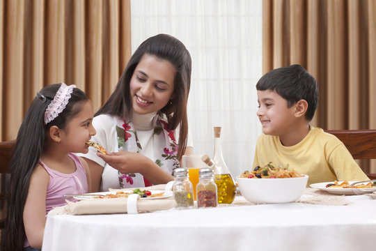 Cheerful mother feeding her daughter pizza with son sitting besides 