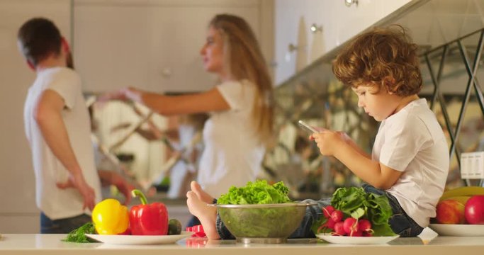 The background of the arguing parents, the mother is slapping the father while the little kid with mobile phone in his hands is sitting on the kitchen furniture in the front.