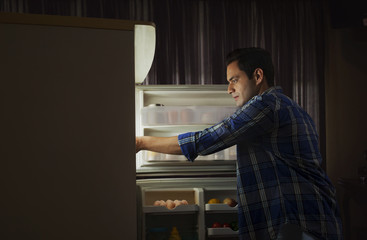 Young man standing in the kitchen searching food in the fridge at night