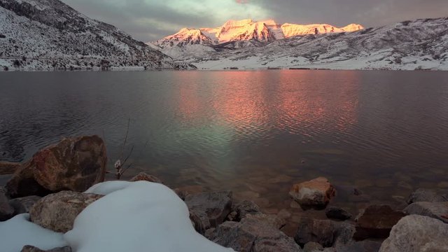 View of snowy landscape reflecting through reservoir at sunrise as sun lights up mountain peaks.