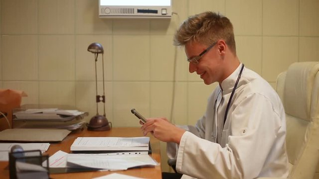 Doctor texting on smartphone while sitting in the office and smiling to the camera

