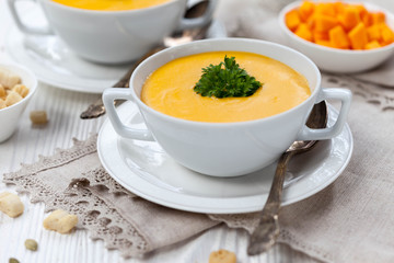 Pumpkin cream soup with croutons, raw fresh pumpkin pieces  and herbs on a white rustic wooden background