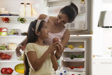 Mother giving apple to daughter in front of open refrigerator